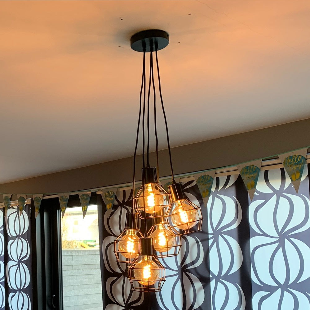 Industrielle Pendelleuchte mit sechs warmlichtfarbenen Glühlampen in einem Metalldrahtrahmen, von unten gesehen und leicht angewinkelt von der Seite, an der Decke in einem modernen Interieur montiert.