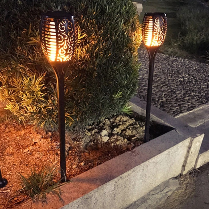Dekorative Solar-Gartenfackeln mit Flammeneffekt, in der Dämmerung entlang eines Gartenweges platziert, von vorne mit leuchtenden Mustern und warmer Stimmungsbeleuchtung.