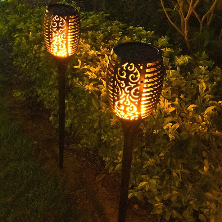 Dekorative Gartenfackeln mit warmem LED-Flammeneffekt, nachts entlang eines Gartenwegs platziert, mit Blick auf die von Sträuchern umgebenen Seiten der Lampen.