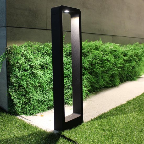 Eine moderne schwarze LED-Gartenlampe mit rechteckigem, offenem Rahmendesign, die auf einem Rasen neben einer Hecke platziert ist; von vorne gesehen, wenn sie nachts leuchtet.