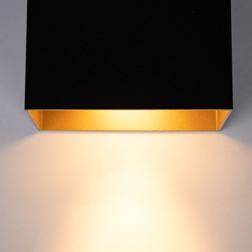 Nahaufnahme einer modernen schwarzen Wandlampe mit goldener Innenseite von unten, die warmweißes Licht nach unten abgibt.