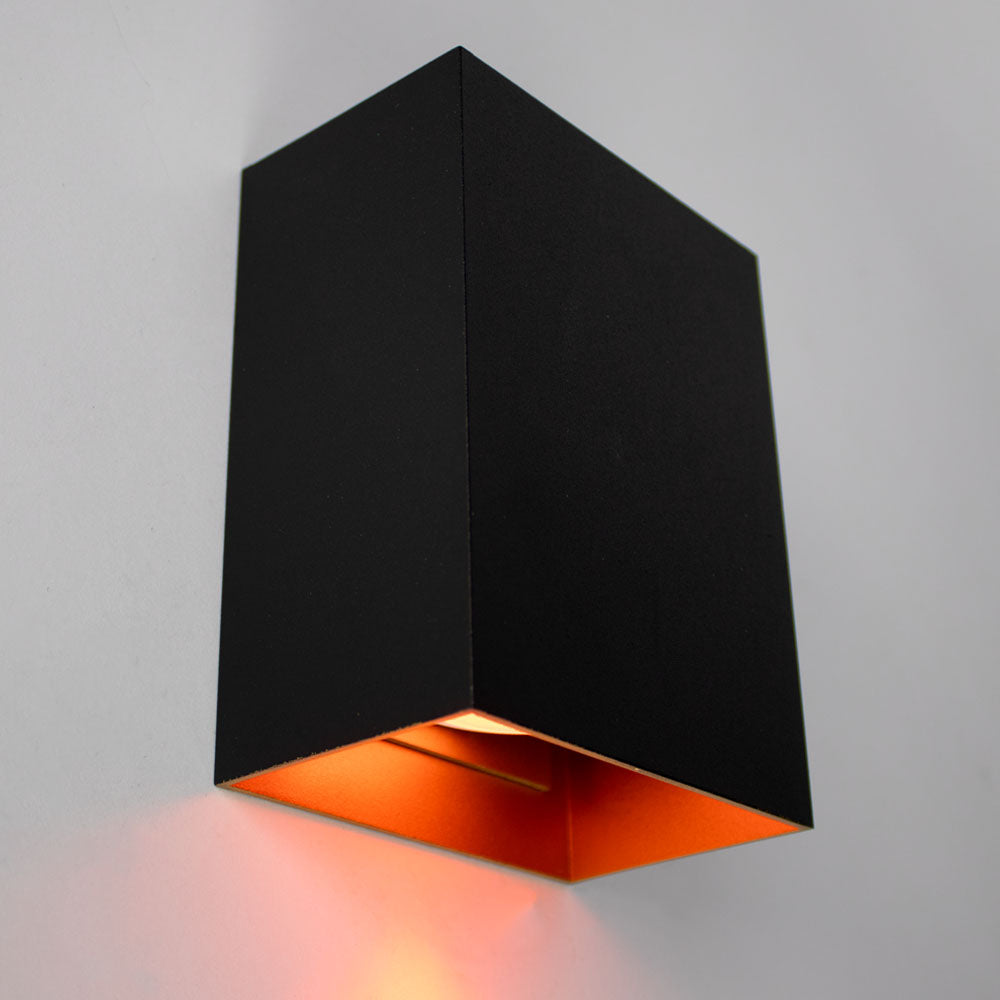 Schwarze moderne Wandleuchte mit rechteckigem Design und kupferfarbener Innenseite, schräg von unten vor einer weißen Wand fotografiert.