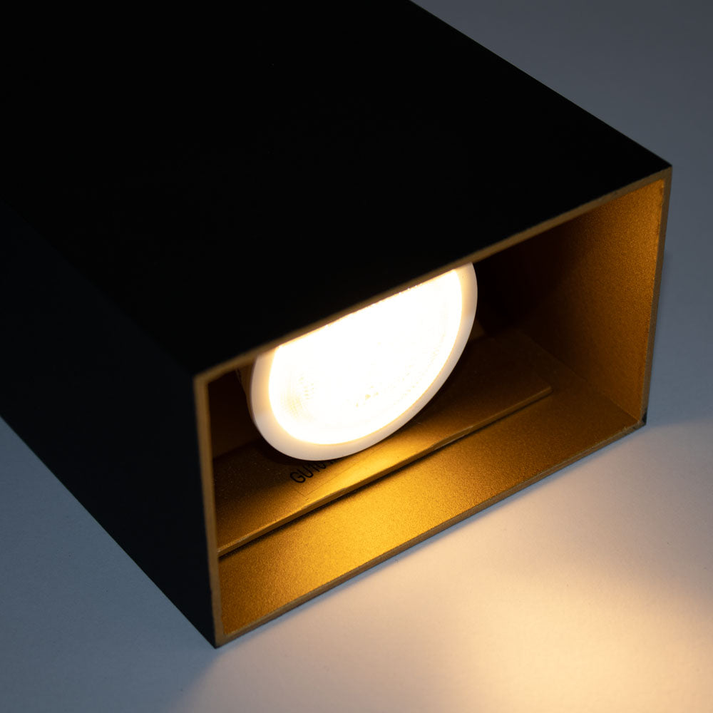 Eine Nahaufnahme von der Seite eines modernen schwarzen Deckenstrahlers in Würfelform mit warmweißem Licht, der die offene Vorderseite mit einem goldfarbenen Inneren für zusätzliche Reflexion zeigt.