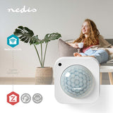 Nahaufnahme des Nedis SmartLife ZigBee-Bewegungssensors mit einem Erfassungswinkel von 120° von vorne, gezeigt in einer modernen Wohnzimmerumgebung mit einer entspannten Frau im Hintergrund.