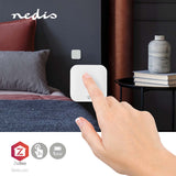 Nahaufnahme einer Hand, die einen weißen intelligenten drahtlosen ZigBee-Wandschalter von Nedis bedient, der an der Wand neben einem modernen Bett in einem stilvoll eingerichteten Schlafzimmer montiert ist – Vorderansicht mit Fokus auf die Produktverwendung.