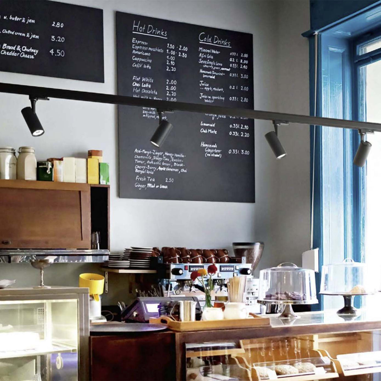 Schienenbeleuchtung mit schwarzen Strahlern an der Decke einer modernen Kaffeebar, Frontalansicht der Beleuchtungsanlage über Theke und Kaffeemaschinen.