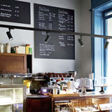 Schienenbeleuchtung mit schwarzen Strahlern an der Decke einer modernen Kaffeebar, Frontalansicht der Beleuchtungsanlage über Theke und Kaffeemaschinen.